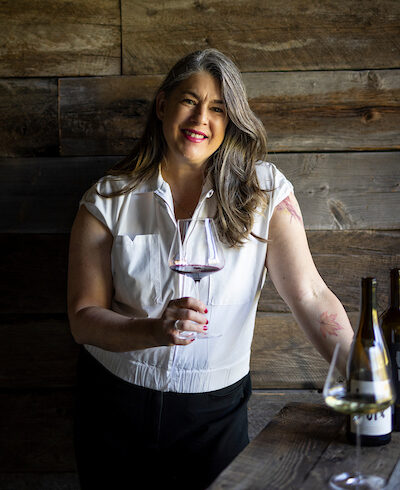 Jaime holding a glass of red wine standing in front of a wood wall.